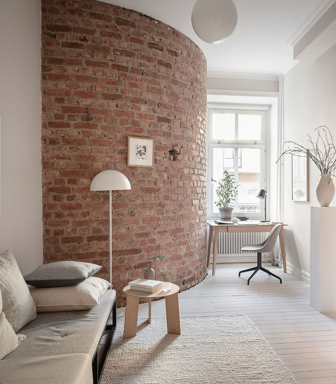 Salón con pared curva de ladrillo visto, sofá gris, mesa auxiliar de madera y escritorio junto a la ventana